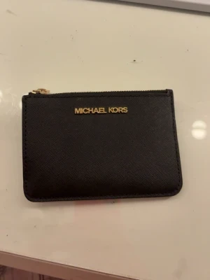 Svart plånbok från Michael Kors - Snygg och stilren svart plånbok från Michael Kors med guldfärgad dragkedja och logga framtill. Tillverkad i tåligt skinnmaterial. Bra skick förutom de på sista bilden. 