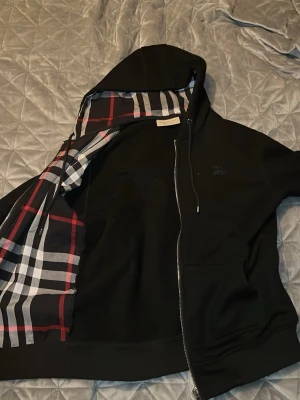 Svart hoodie från Burberry med dragkedja - Cool svart hoodie från Burberry med klassiskt rutigt foder i huvan och insidan. Hoodien har dragkedja framtill, fickor och snören vid huvan. Perfekt för dig som vill ha en stilren och exklusiv look. Materialet är mjukt och bekvämt. Stl M Skriv vid frågor. Inte äkta