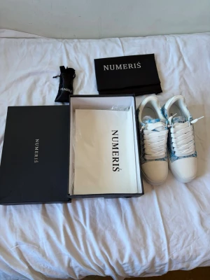 Numeris blåvita sneakers med mönster - Säljer ett par snygga sneakers från Numeris i vitt och ljusblått med coolt mönster. Skorna har vita snören, grov sula och detaljer i textil och läderimitation. Perfekta för dig som vill sticka ut med en unik look.