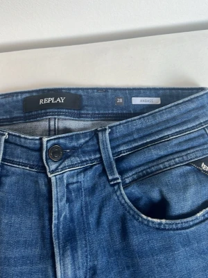 Blå jeans från Replay Anbass - Säljer ett par blå jeans från Replay, modell Anbass. Klassisk femficksdesign med knappgylf och snygga slitningar. Jeansen har en smal passform och är tillverkade i stretchigt denimtyg för extra komfort. Perfekta för en trendig och avslappnad look.