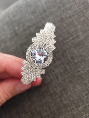 Glittrigt armband med stor kristall - Snyggt armband i silverfärg med massor av små gnistrande stenar och en stor rund kristall i mitten. Armbandet har en unik geometrisk design som verkligen sticker ut och ger en lyxig känsla. Perfekt för dig som vill ha något som syns och glänser extra mycket.