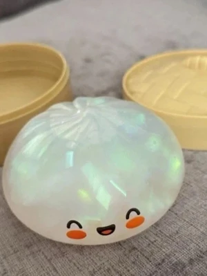 Äkta Glitter dumpling för barn - En liten burk i form av en dumpling med pärlemorskimrande yta och ett gulligt ansikte med röda kinder. Perfekt för att förvara småsaker inom skönhet, som smycken eller hårnålar. Locket är beige och påminner om en bambukorg.