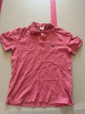 Rosa pikétröja från Lacoste - Säljer en rosa pikétröja från Lacoste i slim fit-modell. Tröjan har klassisk krage, korta ärmar och två knappar framtill. Ikoniska Lacoste-loggan med krokodil på bröstet. Perfekt för en avslappnad och sportig stil.