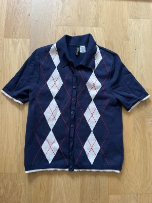 Mörkblå argyle stickad kortärmad tröja - Snygg mörkblå stickad tröja från HM med klassiskt argylemönster i vitt och röda detaljer. Tröjan har korta ärmar, krage och knappar hela vägen framtill. Perfekt för en preppy look och passar till många olika outfits.