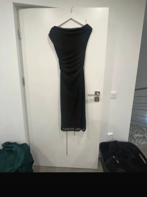Svart långklänning  - Elegant svart långklänning med draperad effekt över byst och midja. Klänningen har en smal passform och är axelbandslös, vilket ger en stilren och modern look. Perfekt för dig som vill ha en klassisk och tidlös outfit.