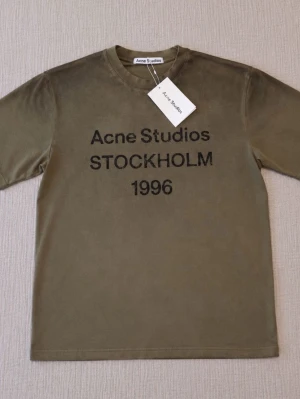 Olivgrön Acne Studios t-shirt - Cool olivgrön t-shirt från Acne Studios med svart text 'Acne Studios STOCKHOLM 1996' på bröstet. Klassisk passform med rund hals och korta ärmar. Perfekt för dig som gillar stilrena och trendiga plagg.