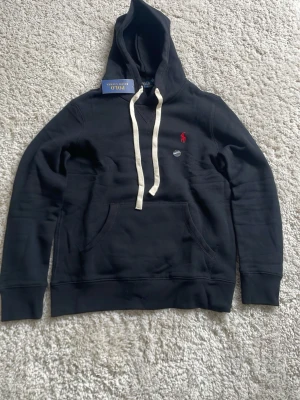 Svart hoodie från Polo Ralph Lauren - Svart hoodie från Polo Ralph Lauren med den klassiska röda logan. Materialet är mjukt och bekvämt, perfekt för en avslappnad look. Den är storlek S men sitter som xs