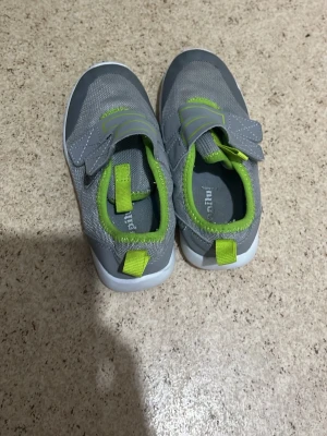 Grå sneakers med gröna detaljer - Säljer ett par grå sneakers från Puma med gröna detaljer och vita sulor. Skorna har meshmaterial och praktiska kardborreband för enkel användning. Perfekt för dig som vill ha ett sportigt och bekvämt alternativ.