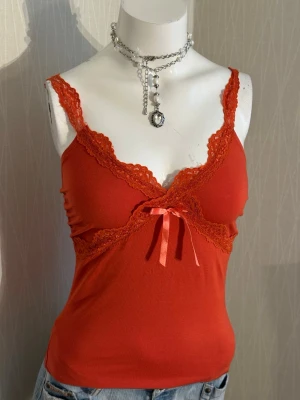 Coral spetslinnetopp med rosett - Snygg coral linnetopp med spetsdetaljer längs urringningen och axelbanden. Toppen har en söt liten rosett framtill och är figurnära i modellen. Perfekt att styla med jeans eller kjol för en trendig look.