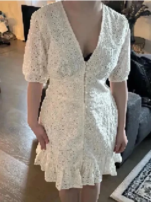 Studentklänning❤️ - Superfin vit kort klänning med broderad spets, v-ringning och knappar framtill. Klänningen har korta puffärmar och volang nertill för en söt och somrig look. Perfekt för varma dagar och midsommar. Liknar Adoore klänningen