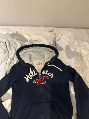 Marinblå hoodie från Hollister - marinblå hoodie från Hollister med dragkedja. Fodrad huva med vitt teddyfoder och vita dragsnören. Perfekt för dig som gillar klassisk college-stil och vill ha något riktigt bekvämt. Jättebra skick och pris kan diskuteras 