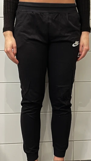 Svarta mjukisbyxor - Snygga svarta mjukisbyxor från Nike med klassisk logga på vänster lår. Byxorna har en smal passform, resår i midjan och muddar vid bensluten. Perfekta för chill eller träning, tillverkade i mjukt bomullsmaterial.