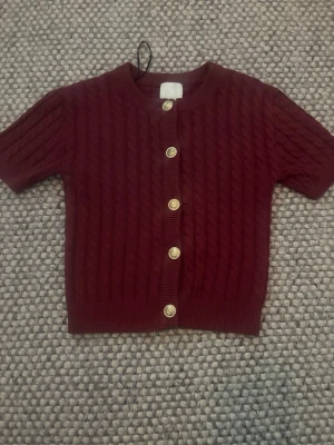 Vinröd kabelstickad kofta kort ärm - Kortärmad vinröd kofta med kabelstickat mönster och fem guldfärgade knappar framtill. Rund halsringning och ribbade muddar vid ärm och nederkant. Perfekt för lager på lager och en snygg detalj till din outfit.