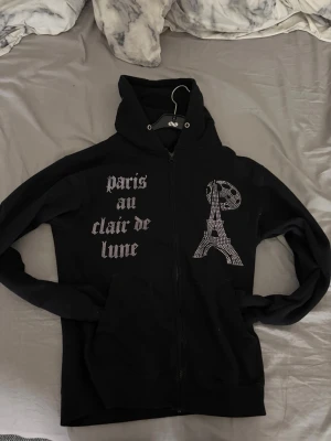 Svart bling lucid  hoodie med Paris-tryck - Säljer en svart hoodie från bling lucid i storlek S med dragkedja och huva. På framsidan finns texten 'paris au clair de lune' i glittriga stenar samt ett motiv av Eiffeltornet. Hoodien har fickor framtill och ribbade muddar. Perfekt för dig som gillar Paris-vibbar och snygga detaljer.
