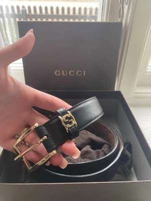 Svart skärp från Gucci i skinn - Snyggt svart skärp från Gucci i äkta skinn med guldfärgat spänne och GG-detalj. Klassisk design som passar till många olika outfits. Levereras med originalbox och dustbag.