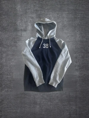 Marinblå hoodie från HM med grå ärmar - Snygg hoodie i marinblått med kontrasterande ljusgrå ärmar och huva från HM. Framtill finns ett tryckt nummer '38' i vitt. Hoodien har dragsko i huvan och muddar vid ärmslut och nederkant. Perfekt för en avslappnad och sportig stil.