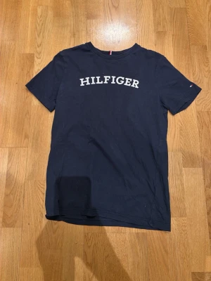 Mörkblå Hilfiger t-shirt - Snygg mörkblå t-shirt från Tommy Hilfiger med klassisk logga tryckt i vitt över bröstet. T-shirten har rund halsringning och korta ärmar. Tillverkad i mjuk bomull som känns skön mot huden. Perfekt för en avslappnad och stilren look.