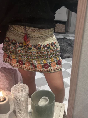 Broderad kortkjol med tofsar - Säljer en superfin kortkjol i beige med färgglada broderade blommor och speglar. Kjolen har en bred resår i midjan och dekorativa tofsar i rött och gult framtill. Perfekt för dig som gillar boho-stil och detaljer.
