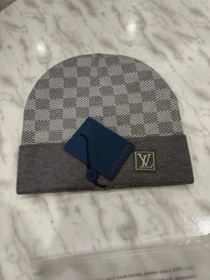 Grå stickad mössa  - En grå stickad mössa med rutigt mönster och uppvikt kant, med en liten logotyp framtill och etikett kvar.