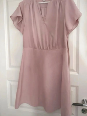 Ljusrosa kort klänning med v-ringning - Söt ljusrosa klänning med kort ärm och v-ringad hals. Klänningen har en avslappnad passform och är sydd i ett mjukt, lätt tyg som passar perfekt till varma dagar. Enkel och stilren design utan mönster eller extra detaljer.
