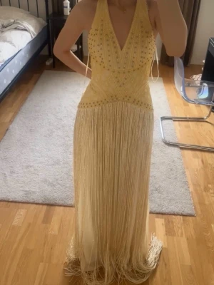 Lång ljusgul fransklänning med pärlor - Säljer en lång, ljusgul klänning med djup v-ringning och halterneck. Klänningen är dekorerad med pärlor upptill och har långa fransar från midjan och ner. Perfekt för dig som vill sticka ut med en unik och glamorös look🤍 Nypris 6800kr