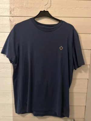 Morris T-shirt storlek S - En stilren blå t-shirt med rund hals och korta ärmar. På bröstet sitter en liten patch med vit och svart detalj. Perfekt basplagg för en avslappnad look.