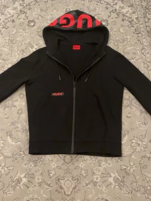 HUGO Boss Zip Hoodie – XS, Nyskick - Säljer en stilren och modern zip hoodie från HUGO Boss. Den svarta designen med röda detaljer ger ett sportigt och exklusivt intryck. Perfekt för våren och svalare sommarkvällar.  • Märke: HUGO (Boss) • Färg: Svart med röda detaljer • Storlek: XS (passar S) • Skick: Som ny, inga skador eller tecken på slitage  Kan hämtas upp eller fraktas.   Hör gärna av dig vid intresse eller frågor!