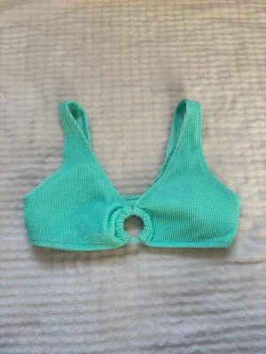 Turkos/grön bikiniöverdel med ringdetalj - Trendig turkos/grön bikiniöverdel i mjukt, strukturerat tyg med en rund ringdetalj framtill. Breda axelband och utan synliga knäppningar för en enkel och bekväm look. Perfekt för strandhäng eller poolparty.