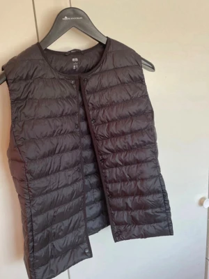Svart dunväst från Uniqlo - Snygg svart dunväst från Uniqlo med quiltad design och tryckknappar framtill. Västen är lätt och smidig, perfekt att slänga över en hoodie eller tröja. Tillverkad i tunt, glansigt material som ger en clean look.