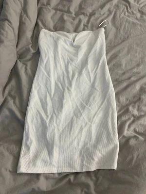 Vit ribbad kort klänning H&M - Säljer en vit ribbad kort klänning från H&M. Klänningen är axelbandslös med en liten v-ringning framtill och har en tight passform. Perfekt för sommarens alla tillfällen och enkel att styla med accessoarer.