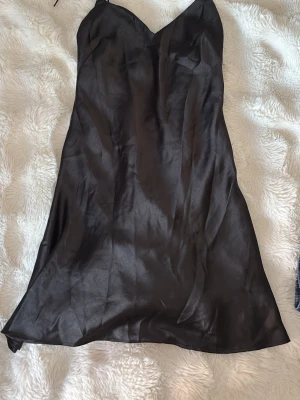 Svart satin slipklänning - Svart slipklänning i satin med v-ringad hals och smala axelband. Klänningen har en slät och glansig yta som ger en elegant look. Perfekt för dig som gillar minimalistisk stil och vill ha något enkelt men snyggt. Man knyter klänningen där bak.