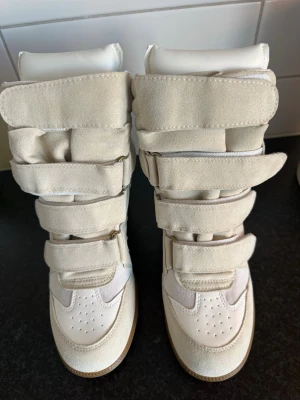 Beige höga sneakers med kardborre - Snygga höga sneakers i beige mocka och vitt skinn med flera breda kardborreband och brun sula. Skorna har en chunky siluett och vadderad design som ger en cool och modern look. Perfekta för dig som vill sticka ut med din stil.