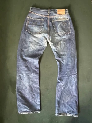 SKIT FETA MOMONTARO  - Säljer ett par blå jeans med unika mönstrade detaljer på bakfickorna och klassisk läderpatch i midjan. Jeansen har rak passform och tydliga slitningar för en cool, avslappnad look. Perfekta för dig som gillar streetwear och vill sticka ut lite extra.