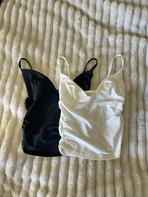 Svart och vit linnetopp med smala band - Två stilrena linnetoppar i svart och vitt med smala axelband och v-ringad hals. Topparna har lätt rynkade sidor för en snygg passform och är perfekta att matcha med jeans eller kjol. Materialet känns mjukt och stretchigt.