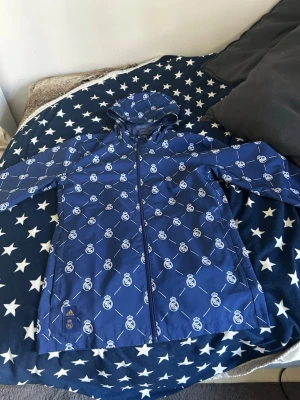 Blå Real Madrid regnjacka Adidas - Snygg blå regnjacka från Adidas med Real Madrid-logga över hela jackan. Jackan har huva, hel dragkedja framtill och är tillverkad i ett vattenavvisande material. Perfekt för regniga dagar och för dig som älskar fotboll och vill sticka ut.