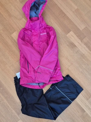 Rosa regnjacka och svarta överdragsbyxor från Tenson - Rosa regnjacka från Tenson med huva och reflexdetaljer. Jackan har dragkedja framtill, två sidofickor och justerbara ärmslut. Jackan kommer i set tillsammans med ett par svarta överdragsbyxor. Materialet är vattenavvisande och perfekt för regniga dagar. Märkt med MPC-teknologi för extra skydd. 