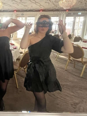 Svart oneshoulder klänning med knyt - Säljer en svart oneshoulder klänning med volangdetaljer och knyt i midjan. Det finns dragkedja i midjan och bandet går att ta av och knyta hur man vill! Den är endast använd en gång🤗 Fler bilder kan tas och pris kan diskuteras😇