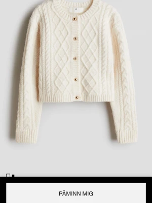 Vit kabelstickad kofta från H&M - Mysig vit kabelstickad kofta från H&M med dekorativa guldknappar framtill. Koftan har ett klassiskt mönster med flätor och rutor, ribbade muddar och rund hals. Perfekt för lager på lager och en stilren look.