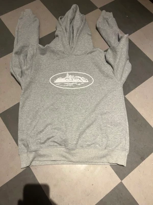 Grå hoodie med tryck framtill - Snygg grå hoodie med huva och ett vitt tryck framtill som föreställer en byggnad eller landskap i en oval ram. Hoodien har klassisk passform, ribbade muddar och magficka. Perfekt för en chill och avslappnad stil.