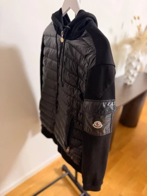 Svart Moncler cardiganjacka  - Snygg svart pufferjacka från Moncler med huva och dragkedja framtill. Jackan har quiltad framsida i glansigt material, mjuka svarta ärmar och fickor med dragkedja. Moncler-logga på ärmen och ribbade muddar. Perfekt för kalla dagar storlek XL