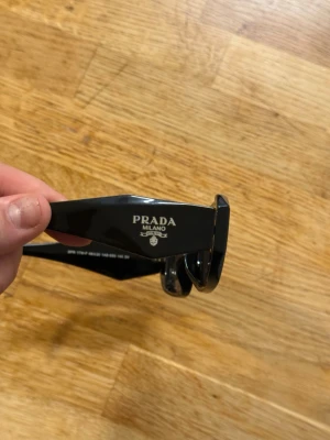 Svarta solglasögon från Prada - Stilrena svarta solglasögon från Prada med rektangulär form och breda skalmar där loggan syns tydligt. Glasögonen har en modern vibe och är tillverkade i blankt syntetmaterial.