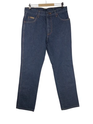 Wrangler Texas blåa jeans, W33 L32 - Ett par Wrangler Texas jeans i en klassisk mörkblå denimfärg. Jeansen har en traditionell design med fem fickor, inklusive en liten myntficka framtill och två bakfickor med Wranglers signatur "W" sömmar. Gylfen stängs med dragkedja och knapp. Det finns en Wrangler-logotyp i läderimitation på bakfickan och en mindre tygetikett på myntfickan. Midja: 41cm Långt: 79cm