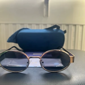 Guldiga ovala solglasögon från Gucci - Snygga solglasögon från Gucci med guldfärgad metallbåge och ovala, mörka linser. Klassisk och stilren design med svarta skalmar och diskret Gucci-logga på linsen. Perfekta för dig som vill ha en exklusiv accessoar med lyxig känsla.