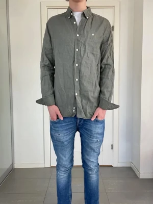 Oliv grön långärmad skjorta från Morris - Säljer en oliv grön skjorta från Morris med klassisk button-down krage och vita knappar. Skjortan har lång ärm och diskret logotyp på bröstet. Perfekt för dig som vill ha en stilren och enkel look.