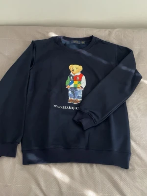 Mörkblå sweatshirt Polo Bear - Cool mörkblå sweatshirt från Ralph Lauren med Polo Bear-motiv på bröstet. Tröjan har rund hals och ribbade muddar. Materialet är mjukt och bekvämt, perfekt för en avslappnad look. Motiv med björn i färgglad outfit och texten 'POLO BEAR by RALPH LAUREN'.