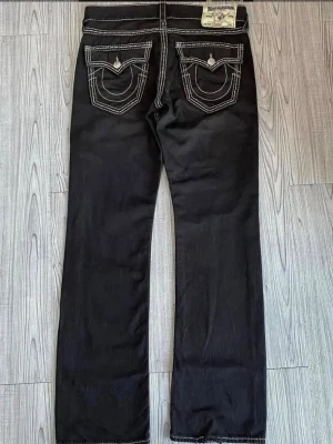 True Religion svarta jeans med kontrastsöm - Svarta jeans från True Religion med raka ben och tydliga vita kontrastsömmar. Bakfickorna har märkets ikoniska broderi och knappar. Klassisk denimlook med coola detaljer, perfekt för dig som vill sticka ut. W34 L32
