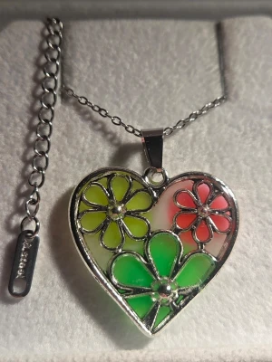Glow Halsband med hjärthänge och blommor - Unikt handgjort halsband med ett hjärtformat hänge i metall, dekorerat med färgglada blommor i grönt, gult och rosa. Hänget har en cool glow-in-the-dark effekt som lyser i mörkret. Kedjan är silverfärgad i rostfritt stål och har justerbar längd. Perfekt för dig som gillar färg och statement-smycken. Både kedjan & hänget tål vatten utan att missfärgas.