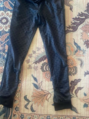 Svarta mjukisbyxor från Adidas - Snygga svarta mjukisbyxor från Adidas med diskret mönster över hela byxan. Byxorna har resår i midjan och muddar vid bensluten, vilket ger en skön passform. Perfekta för chill eller träning.