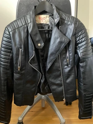 Svart skinnjacka från Vojelles - Cool svart fuskskinnjacka från Vojelles med snygga detaljer som quiltade axlar och ärmar, silverfärgade dragkedjor. Jackan har två vertikala bröstfickor med dragkedja och en asymmetrisk front. Den har tecken på användning runt nacken som syns i bilderna 