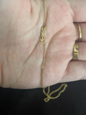 Guldhalsband med YSL-logo - Snyggt och stilrent halsband i guldton med den ikoniska YSL-logotypen som hänge. Kedjan är tunn och elegant, perfekt för att lyfta vilken outfit som helst. Materialet ser ut att vara metall med guldfinish och logotypen är i samma färg.
